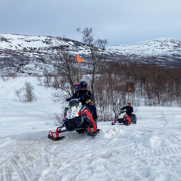 Två röd-vita snöskotrar i fjällmiljö kommer körande mot kameran