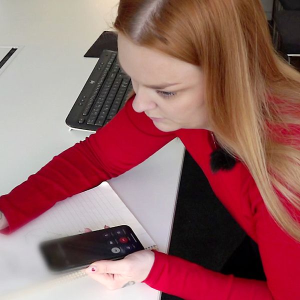 SVT:s reporter Anna Wallbrandt sitter med pappaer och penna och pratar i telefon med högtalare på. Hon har en röd tröja på sig och rödlätt hår.