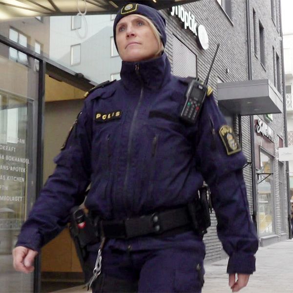 En polis i uniform, Therese Forslund, går längs en gata i Kiruna centrum. Flera byggnader syns i bakgrunden. I nära anslutning finns en entré till en butik, och längre bort syns en traktor gräva i en gata.
