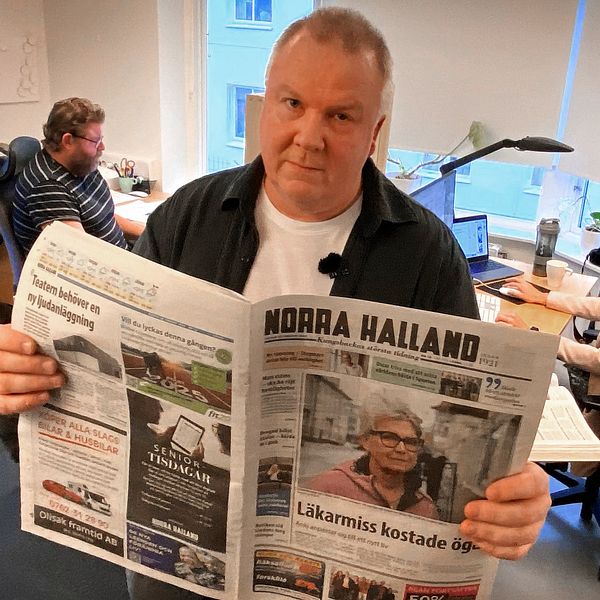 En person håller upp en tidning med titeln ”Norra Halland” i ett kontorsutrymme där två andra personer arbetar vid skrivbord. Tidningsrubrikerna är synliga, och rummet har naturligt ljus som strömmar in från ett fönster.