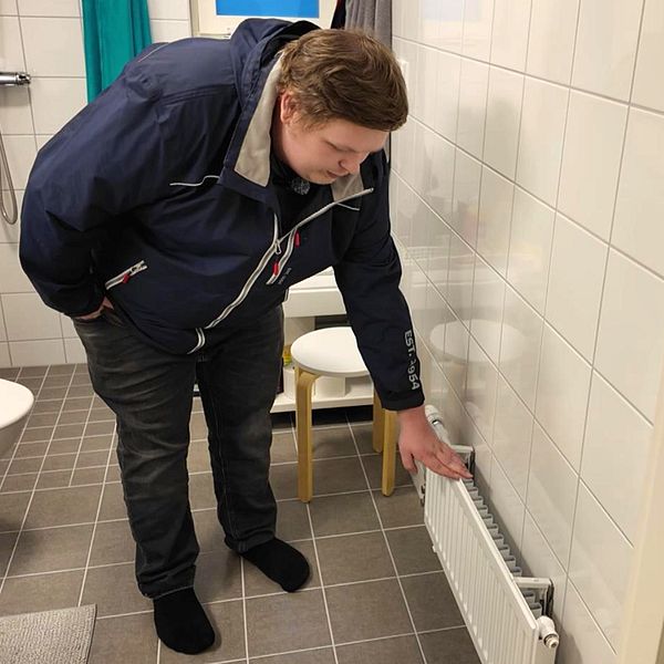 Jonatan Ståhl står i sitt badrum och pekar på en radiator längs väggen. I bakgrunden syns en toalett och en stol. Badrummet har ljusa väggar och en grå keramikplattläggning på golvet.