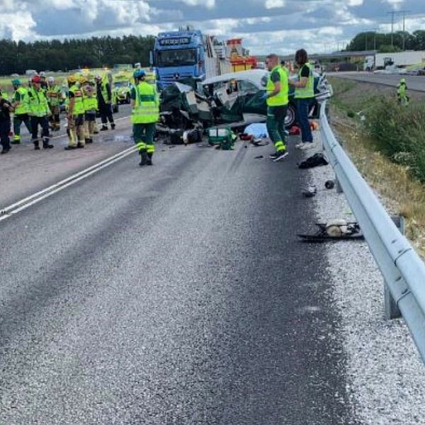 En trafikolycka på en motorväg med flera räddningspersonal som arbetar vid olycksplatsen. Två bilar är involverade, varav en verkar ha skadats allvarligt. Räddningstjänst och ambulanspersonal är synliga, och vägen är avspärrad. I bakgrunden syns trafik.