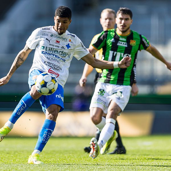 Fotbollsspelare i vit och blå dräkt sparkar bollen, motståndare i grönsvart förföljer.