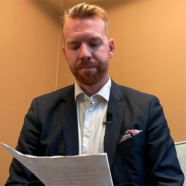 En man i kostym sitter i en soffa och håller i ett papper. Han är fotograferad från en frontvinkel i en inomhusmiljö med väggar i en varm nyans. Det finns en kudde i bakgrunden.
