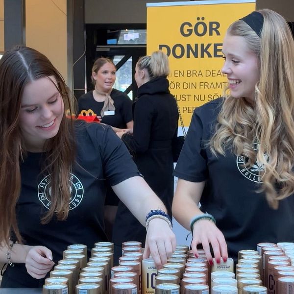 Två personer i svarta tröjor står vid ett bord fyllt med små kartonger eller burkar i en köpcentrummiljö. Bakom dem står skyltar med texten ”Gör Donken!” och information om jobbansökningar hos McDonald's.