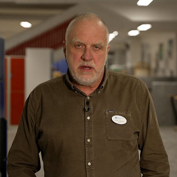 En person i brunt skjorta står framför en skola med blå och röda skåp i bakgrunden, vilket skapar en miljö som antyder en utbildningsinrättning.