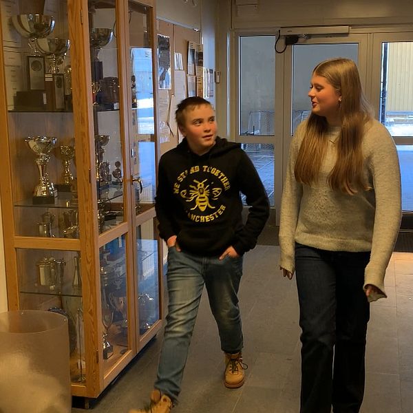 Två personer går genom en korridor i en skola eller idrottsanläggning. I bakgrunden syns en glasmonter innehållande ett flertal pokaler och priser.