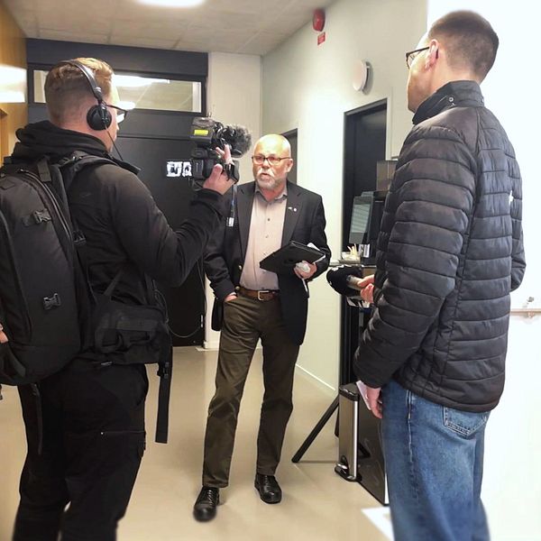 En reporter och en kamerateam står i en korridor, där reportern intervjuar en person som håller i papper. Den tredje personen observerar från sidan. Korridoren har ljusa väggar och dörrar på vardera sidan.