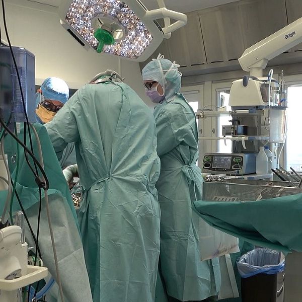 Kirurgisk operation som pågår i ett operationsrum. Flera sjukvårdspersonal i skyddsutrustning arbetar tillsammans kring en patient på operationsbordet. Utrustning och monitors syns i bakgrunden, och det råder en koncentrerad atmosfär.