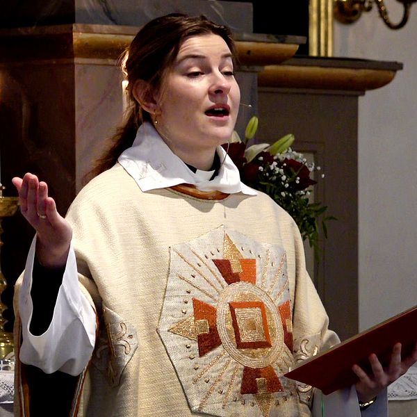 En präst, 24-åriga Evelina Sjöwall, klädd i en ornamenterad mässhake håller en öppen bok och verkar vara mitt i en predikan i en kyrka. En tänd ljusstake syns i bakgrunden.