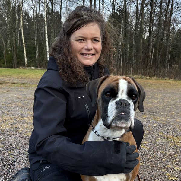 Veronica Frohlund sitter på marken med en boxerhund, omgiven av en skogsliknande miljö med träd i bakgrunden. Personen bär en svart jacka och kramar om hunden med ena armen. Hundens uttryck är vänligt och nyfiket.