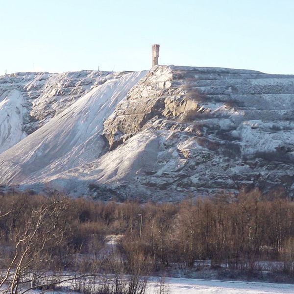 En snötäckt bergsformation med branta sidor, toppad av ett tornliknande struktur, omgiven av ett landskap med snö och trädkronor.