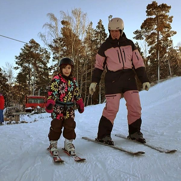 Ett barn och en vuxen står i en skidbacke, bägge har hjälm. Barnet har en blommig overall, mamman en skiddräkt i svart och rosa.