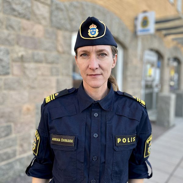 Polis i uniform står på en gata med stenlagda hus i bakgrunden. Uniformen är mörkblå med gula detaljer, och det finns en polishuvudbonad med en emblem på toppen. Några fönster och en byggnad syns i bakgrunden.