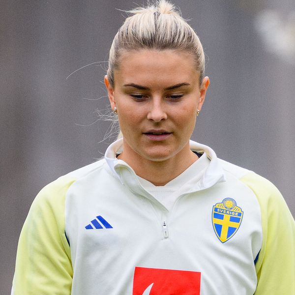 En person klädd i en svensk fotbollströja står framför en suddig bakgrund. Tröjan har ett blått och gult svensk landslagsmärke samt en röd logotyp på bröstet.