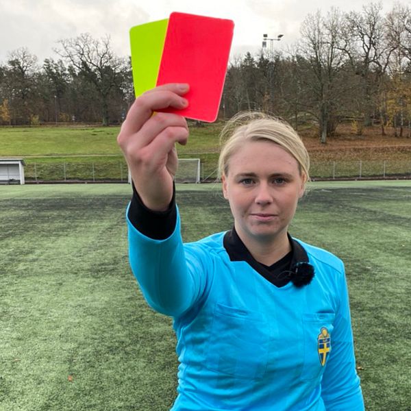 Kvinna i blå domartröja står på fotbollsplan och visar upp ett gult och ett rött kort.