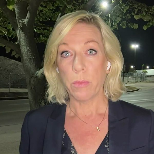 En kvinna (Ginna Lindberg, USA-korrespondent för Sveriges radio) står utomhus i mörker, iförd en mörk blazer. Bakgrunden visar en väg som är svagt upplyst av gatulampor, med träd och en grusig yta i närheten.