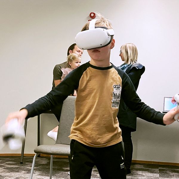 En pojke med VR-headset utforskar en virtuell verklighet medan han står i ett rum. I bakgrunden finns flera personer som observerar honom och ett skrivbord med en dator.