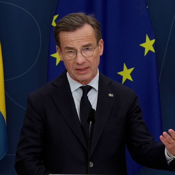 Ulf Kristersson (M) står och pratar vid ett podium framför en blå bakgrund med Sverige- och EU-flaggor i Regeringskansliets pressrum.