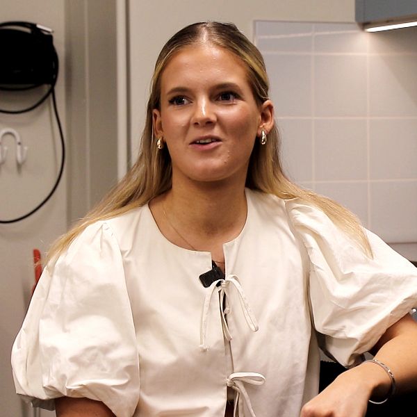 En kvinna med långt hår sitter i en inomhusmiljö i en ljus blus.