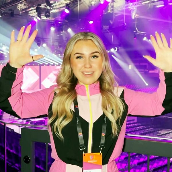 Lia Larsson står i Melodifestival-arenan med rosa kläder och händerna upp i luften