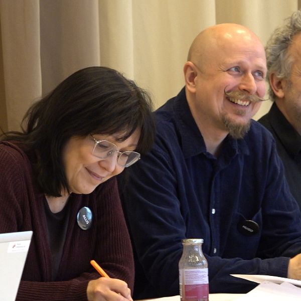Tre personer sitter på rad vid ett bord. Till vänster en kvinna som antecknar något pennan, i mitten en man som tittar uppåt och ler. Till höger en man som tittar upp och ler.