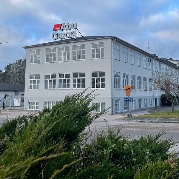 Byggnaden med skylten ”Abu Garcia” i tydliga bokstäver, omgiven av buskar och träd. Det är en grå, fler våningsbyggnad med stora fönster. Himlen är molnig och vädret verkar kallt. En vägskylt och en väg kan ses i förgrunden, vilket indikerar att bilden är tagen nära en väg.
