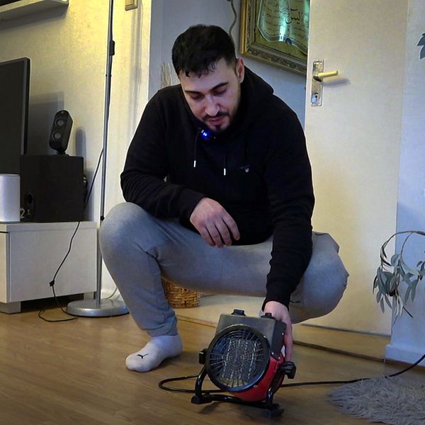 En person i en modern inomhusmiljö, klädd i svart hoodie och gråa byxor, lutar sig framåt för att justera en liten värmefläkt. I bakgrunden syns en stor TV och en grön växt.