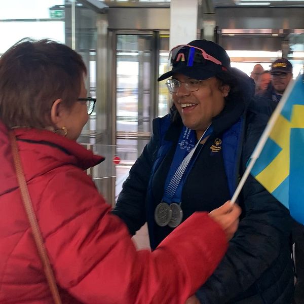 Person i röd jacka överlämnar svensk flagga till medaljprydd individ i vintermiljö.