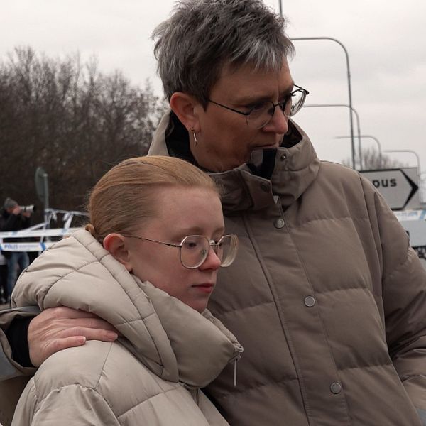 Två personer står utomhus iförda tjocka vinterjackor. Området bakom dem är avspärrat med polistejp och det finns några polisbilar synliga i bakgrunden. Det är en kall vinterdag med snö på marken.