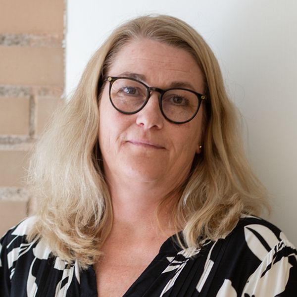 Kvinnlig person med blont, vågigt hår, klädd i en svart och vit blommig blus, står framför en ljus vägg av sten.