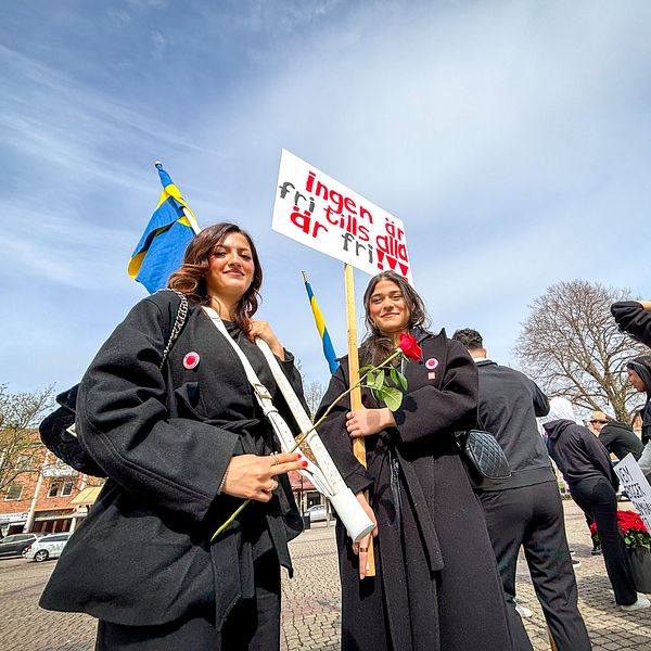 Två kvinnor i svarta kappor bär skylt och rosor vid första maj-demonstration, svenska flaggor i bakgrunden.