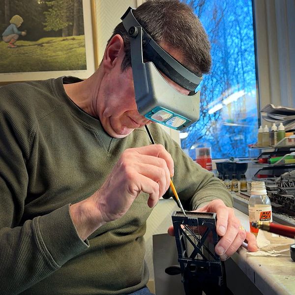 En person sitter vid ett arbetsbord och målar detaljer på en modell med en pensel. Runt dem finns olika verktyg och material för modellbygge, inklusive färgburkar och penslar. I bakgrunden syns en fönsterutsikt samt en målning på väggen av ett barn som leker i naturen.