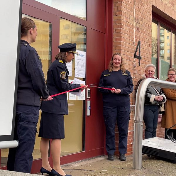 Personer i uniform står vid entrén till en byggnad under en invigning där en kvinna klipper av ett rött band. I bakgrunden finns en skärm och flera personer som applauderar, vilket indikerar att de firar öppnandet av en nyhet eller ett projekt.