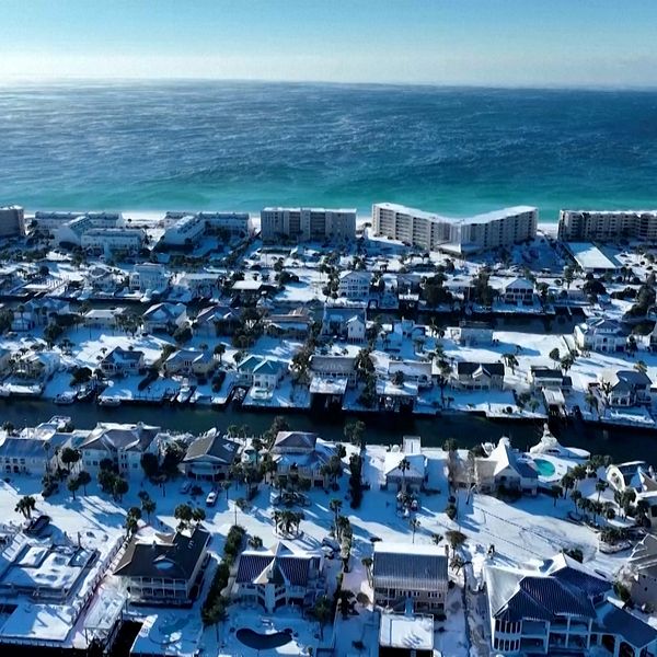 Fågelperspektiv över ett kustområde täckt av snö, där villor och bostadshus i olika storlekar syns. I bakgrunden sträcker sig det blå havet ut mot horisonten, vilket kontrasterar mot den snövita stranden och de snötäckta tak som pryder området.