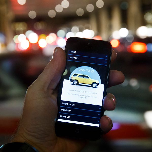Hand håller smartphone med taxi-app, suddig stadsmiljö i bakgrunden.