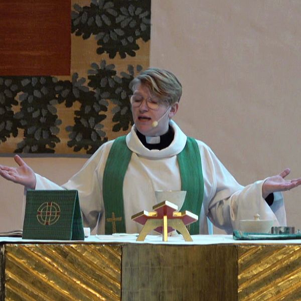 En präst i liturgiska kläder står vid ett altare med uppsträckta armar, flankerad av två tända ljus. På altaret finns en bibel eller religiös bok och ett krucifix eller en religiös symbol. Bakgrunden pryds av dekorativa element.