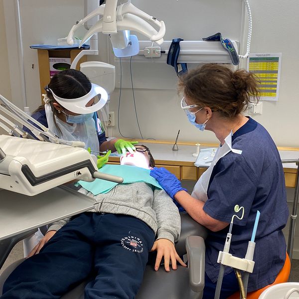 En tandläkarmottagning där en ung patient ligger i en tandläkarstol, omgiven av två tandvårdspersonal som arbetar med behandling. Utrustning inklusive en tandläkarstol och belysning är synlig i bakgrunden.