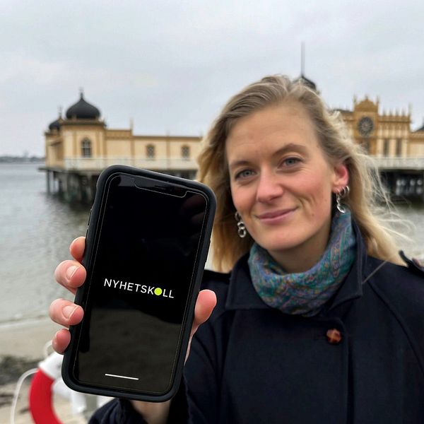 En person står vid en kust med en historisk badhusbyggnad i bakgrunden och håller upp en mobiltelefon som visar appen ”Nyhetsskolan” på skärmen.