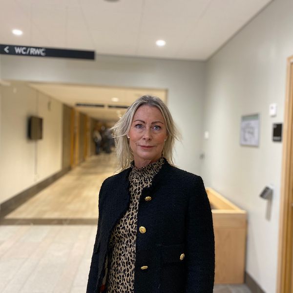 En person står i en korridor med neutrala väggar och belysning. Personen bär en svart kavaj och en blus med leopardmönster. I bakgrunden syns en annan person och en dörr som leder till en toalett, markerad med skyltar.