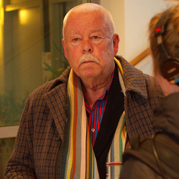 Man med vitt kort hår iklädd en rutig rock och en randig halsduk. Mannen tittar snett förbi kameran. I förgrunden en kvinna med hår i knut och hörlurar, svart jacka.
