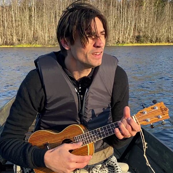 Gustaf Nohrén sitter i en båt på en älv och spelar på en ukulele. Runt omkring finns skog med lövträd, och det är en klar dag med soligt väder. Vattnet speglar omgivningarna.