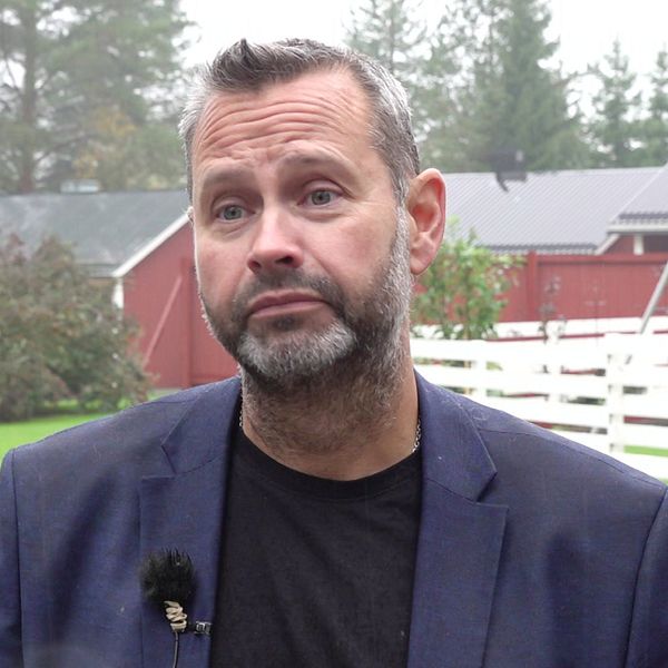 Mattias Riegertzon står framför en grön gräsmatta med träd och en röd byggnad i bakgrunden. Han  bär en mörk jacka och har en mikrofon fäst på kläderna. Det är en dimmig dag, vilket ger en dämpad atmosfär.