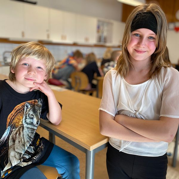 Två elever i klassrum. Vända mot kameran. En pojk som sitter i sin bänk och en flicka som står intill honom.