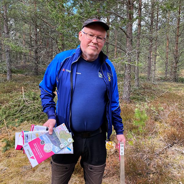 Man i skog håller orienteringskartor och en kontrollstav, omgiven av träd och mossa.