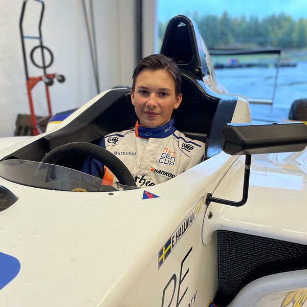 En förare sitter i en racingbil i ett garage. Bilen har en vit lackering med blå och gula detaljer. Bakgrunden visar delar av garaget och ett fönster med utsikt över en grönskande miljö. Föraren bär en overall med sponsormärken och är fastspänd i sätet.