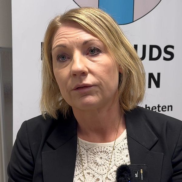 En person i en svart kavaj sitter framför en bakgrund med en logotyp som innehåller blå och rosa element. Personen har på sig en ljus blus och det finns dokument och en skrivbordssak synliga i bakgrunden.