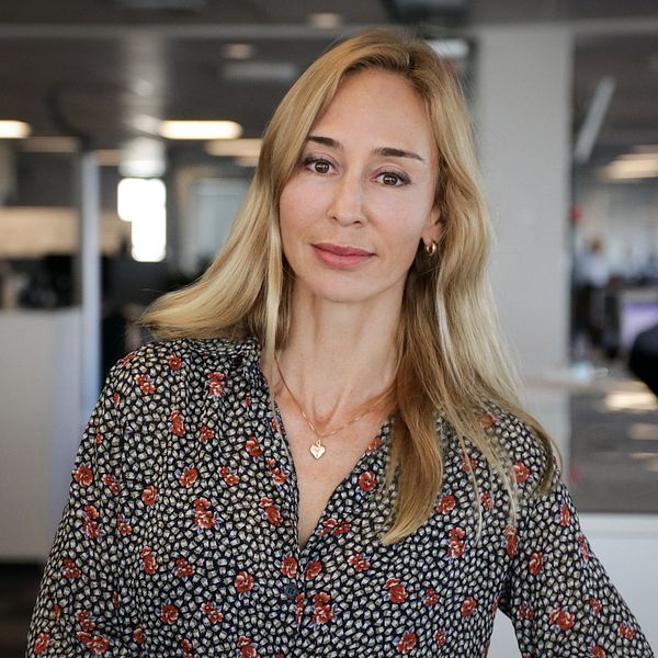 Kvinna med blont hår står på en kontorsarbetsplats med skrivbord och datorer i bakgrunden.
