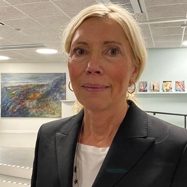 En kvinna i mörk kavaj står i inomhusmiljö med ett konstverk i bakgrunden. Rummet har skarpa, moderna linjer och är inrett med växter samt en hylla fylld med böcker eller tidskrifter på en vägg.