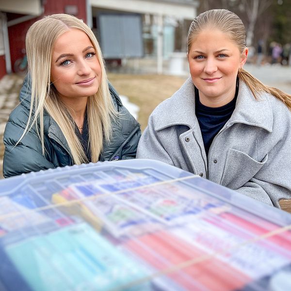 Två unga kvinnor ler bakom en plastlåda fylld med förnödenheter.